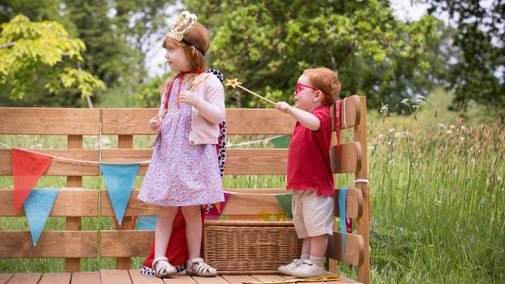 Two young children stand on a wooden outdoor stage holding props.